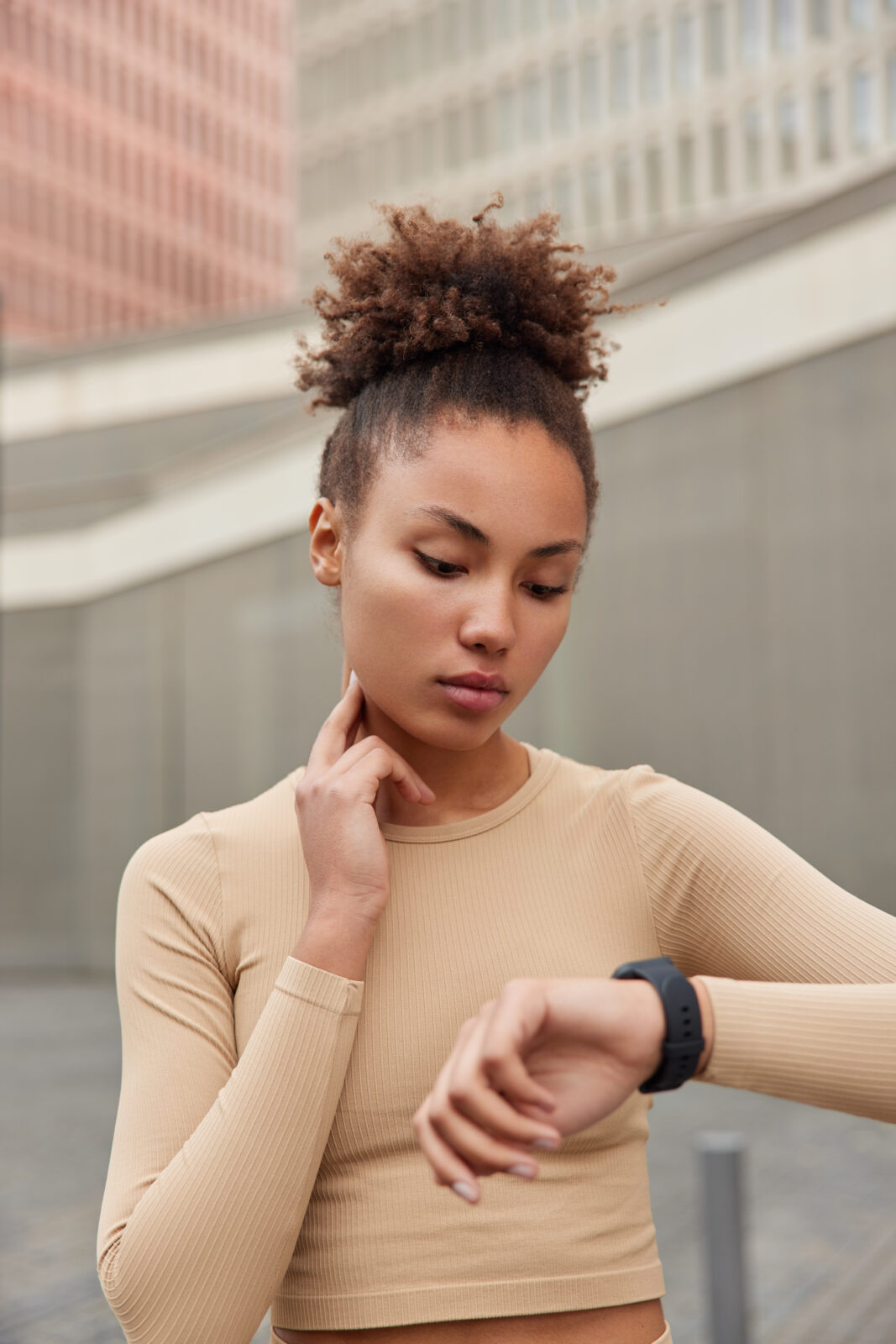 curly haired woman checks pulse on neck focused at smartwatch dressed in sportswear monitors heart rate from wrist poses outdoors.